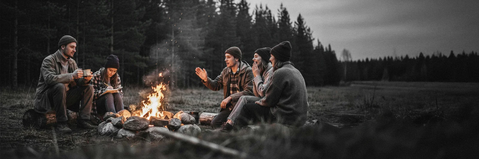 Five people in woollen hats talking and laughing around a campfire in a forest clearing at dusk; one writes in a notebook.